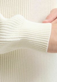 Tessuto a coste di colore crema con una finitura testurizzata. Primo piano del polsino della manica che mostra le coste verticali e un aspetto morbido e lavorato a maglia.