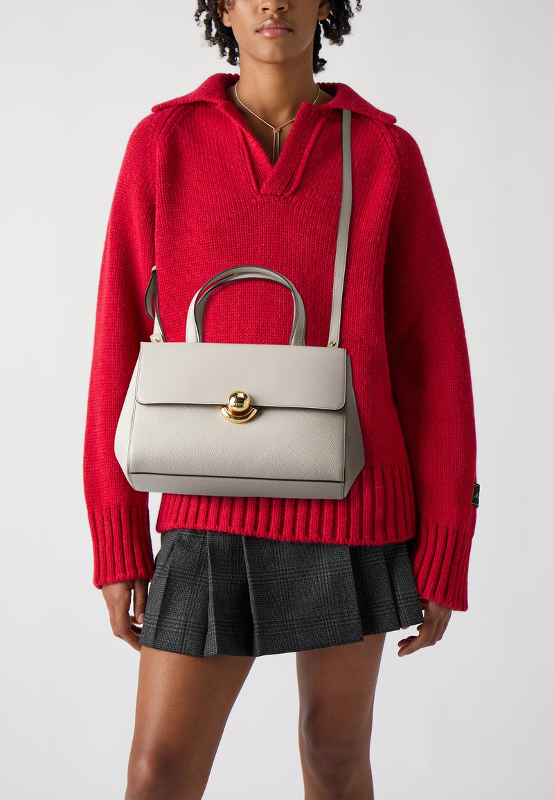 Sac à main en cuir gris à la forme structurée, avec un fermoir en métal doré rond et des poignées courtes. Porté avec un pull en maille rouge et une jupe à carreaux.