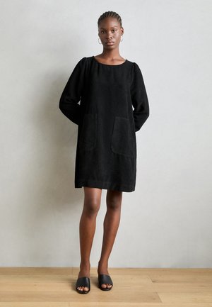 Rochie din corduroy negru cu mâneci lungi, decolteu rotund, două buzunare frontale și o croială lejeră, purtată cu sandale negre tip slide.
