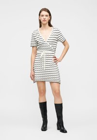 Γυναίκα που στέκεται με το χέρι στη μέση, φορώντας κοντό άσπρο φόρεμα με μαύρες κυματιστές ρίγες και μαύρες μπότες μέχρι το γόνατο.