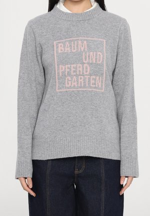 Grijze gebreide trui met geribbelde manchetten en zoom, met roze tekst "BAUM UND PFERD GARTEN" binnen een vierkant op de borst, gedragen over een wit shirt.