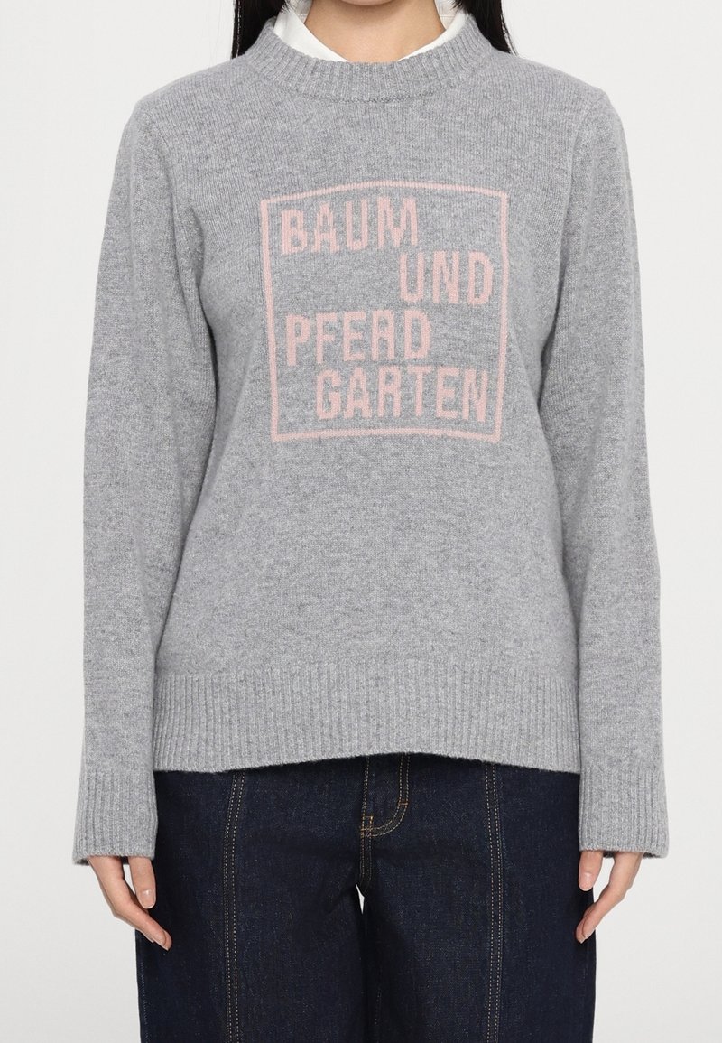 Pull en maille grise avec poignets et ourlet côtelés, arborant un texte rose "BAUM UND PFERD GARTEN" à l'intérieur d'un carré sur la poitrine, porté par-dessus une chemise blanche.