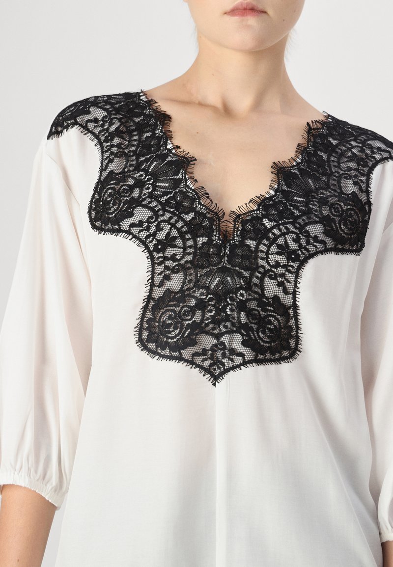 Blouse blanche avec un décolleté en V profond, ornée de délicates bordures en dentelle noire et de détails à franges. Les manches trois-quarts viennent compléter le design.