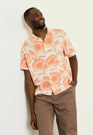 Lächelnder Mann lehnt an einer Wand, trägt ein weißes Kurzarmhemd mit orangefarbenem Sonnenblumendruck und braune Hose, eine Hand in der Tasche.