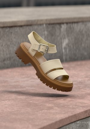 Sandale en cuir beige avec deux brides et une boucle à la cheville, semelle épaisse marron à crampons, positionnée en l'air au-dessus d'une surface en pierre.