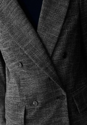 Šedý dvouřadový blazer vyrobený z texturované tkaniny, s kostkovaným vzorem a černými knoflíky. Detailní záběr na klopy a přední stranu.
