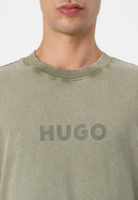 Olive green cotton t-shirt with a ribbed neckline and a large, tonal "HUGO" logo on the chest. Soft texture with a relaxed fit.