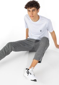 Bílý krátký rukáv atletického trička z měkké síťoviny s černým logem pavouka, spárovaný s šedými zúženými kalhotami se zipy na boku a bílými teniskami.