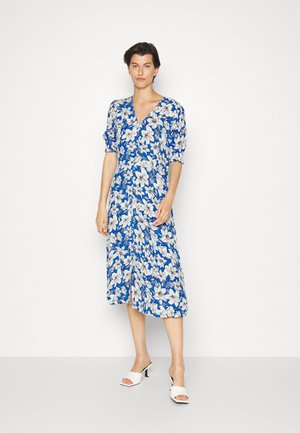Vestido floral azul com flores brancas, decote em V, mangas curtas bufantes, comprimento até ao joelho, tecido leve, combinado com mules brancas de bico aberto.