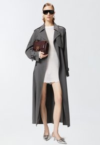 Cappotto grigio con vita cinturata, abito in rete bianca, borsa bordeaux e décolleté argentati a punta. Occhiali da sole neri e orecchini a cerchio completano il look.