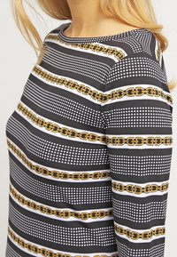 Kvinna som bär en långärmad svart skjorta med vita prickade linjer och horisontella mönster av gyllene kedjor, visad från nacken till midjan.