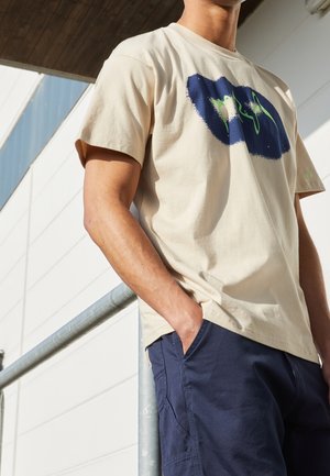 Mężczyzna ubrany w beżowy t-shirt z nadrukiem i granatowe spodnie stoi opierając się jedną ręką na zewnętrznej balustradzie, z ręką w kieszeni.