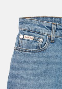 Světle modré džíny z denimu zblízka, ukazující přední kapsu s kovovými nýty a malou nášivkou Calvin Klein umístěnou nad kapsou.