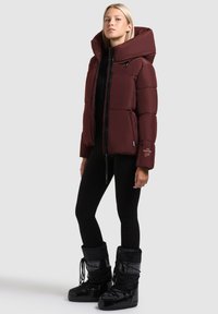 Burgundy puffer jacket with a large hood, zip closure, and side pockets. Paired with black insulated boots featuring lace-up design.