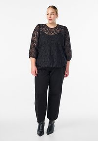 Čierny čipkovaný top s trojštvrťovými rukávmi, okrúhlym výstrihom a voľným strihom, kombinovaný s čiernymi nohavicami a členkovými čižmami s dekoratívnymi cvočkami.