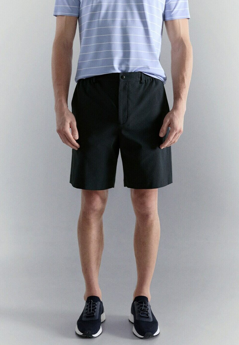 Homme portant un short noir mi-genoux, un polo rayé bleu clair et des baskets noires, debout devant un fond uni.