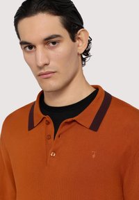 Pomarańczowa koszulka polo z ciemnofioletowym kołnierzykiem, z trzema guzikami z przodu i haftowanym logo. Gładka faktura i dopasowany krój.