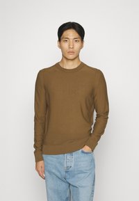Brauner Strickpullover mit rundem Kragen, gerippten Bündchen und Saum. Subtiler Logodruck auf der Brust. Kombiniert mit hellblauen Jeans.