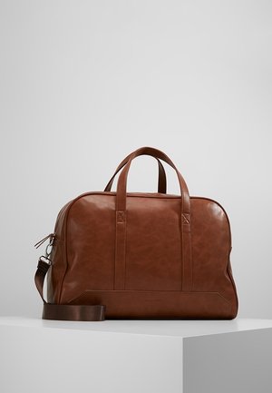 Braune Lederreisetasche mit zwei Griffen und abnehmbarem Schulterriemen, präsentiert auf einem weißen Sockel vor einem hellgrauen Hintergrund.