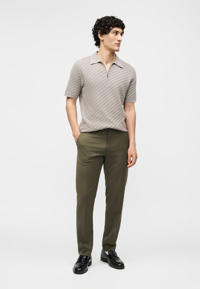 Άνδρας που φοράει ανοιχτό γκρι πικέ πόλο με κοντά μανίκια, χακί παντελόνι και μαύρα δερμάτινα μοκασίνια, όρθιος με τα χέρια στις τσέπες.