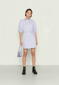 Vestido curto lilás com mangas bufantes, fecho de botão e um cinto de tecido combinante. Combinado com botas de cano baixo pretas e uma pequena mala.