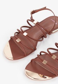 Bruine sandalen met banden en een platte zool, voorzien van gouden vierkante accenten en een metalen neuskap. Glad oppervlak, verstelbare enkelband.