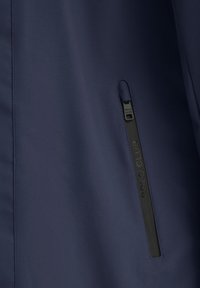 Sezione della giacca navy con una texture liscia, caratterizzata da una tasca con zip e il marchio stampato "POLO CLUB" sul cursore della zip.