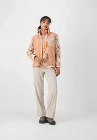Fleece veste maigi oranžā krāsā ar teksturētu rakstu un priekšējo kabatu, saskaņota ar rakstainu garām piedurknēm kreklu un bēšiem biksēm.