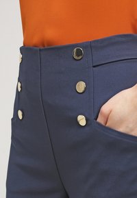 Wysokowysokie granatowe spodnie wykonane z gładkiej tkaniny, ozdobione złotymi guzikami i wyposażone w boczne kieszenie. Pas ma strukturalny fason.