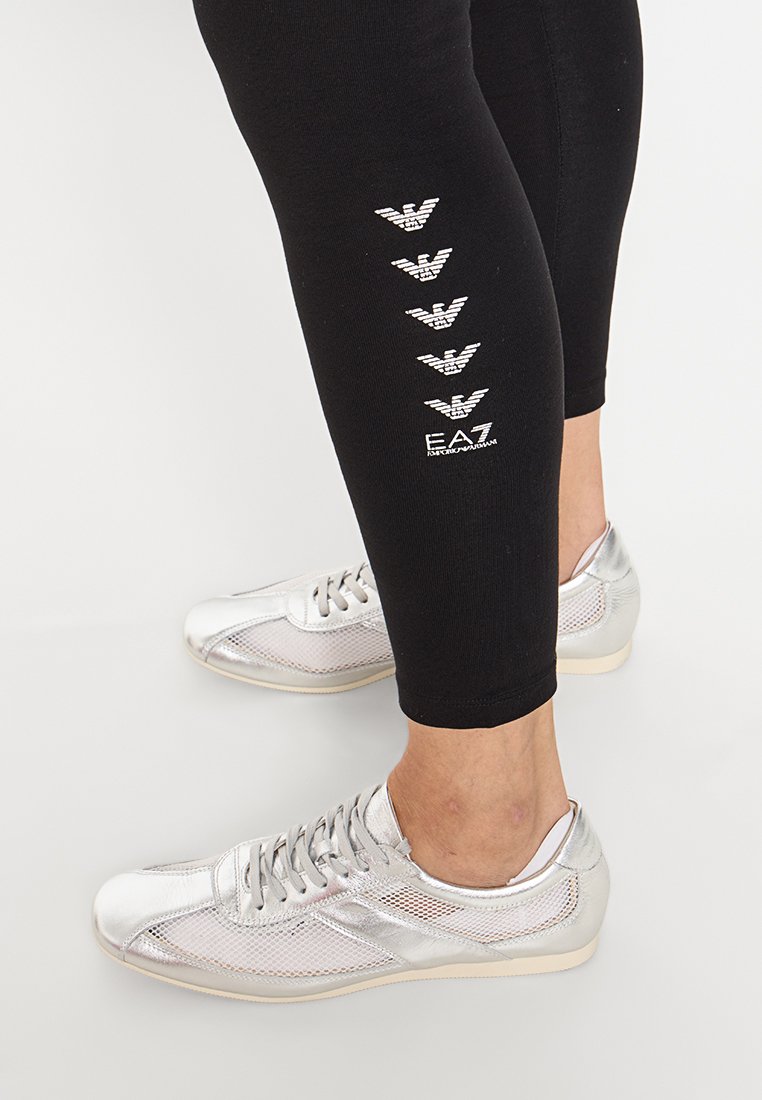 Czarne legginsy z pięcioma białymi logo EA7 po boku, w połączeniu z błyszczącymi srebrnymi sneakersami z siateczkowym wykończeniem i gładką powierzchnią.