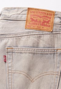 Jean en denim bleu clair avec un patch logo en cuir marron clair. Caractéristiques : surpiqûres orange et petite étiquette rouge sur la couture de la poche.