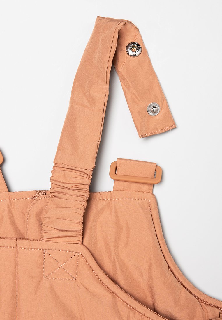 Beige Latzhosen mit verstellbaren Trägern, elastischen Details und Druckknöpfen. Verfügt über eine glatte Textur und minimale Nähte.