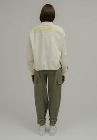 Veste oversize crème avec le texte néon vert "Good Karma Club" dans le dos, associée à un pantalon cargo vert olive et des baskets blanches.