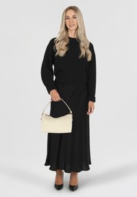 Pull en maille noir associé à une longue jupe noire. Tient un sac à main de couleur crème avec une forme structurée et une poignée incurvée. Talons noirs.