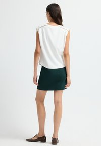 Vrouw die met de rug naar de kijker staat, gekleed in een witte mouwloze top met parel-details en een donkergroene mini-jurk met bruine platte schoenen.