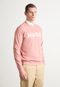Ružová mikina s výrazným bielym logom "HUGO", okrúhlym výstrihom, ribbovanými manžetami a mäkkou tkaninou. V kombinácii s khaki nohavicami.