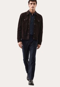 Bruine suède jas, marineblauw patroond blouse, marineblauwe broek en zwarte schoenen. Jas heeft twee borstzakken en metalen knopen.