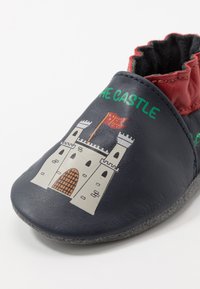 Chaussure en cuir bleu marine avec des accents rouges, présentant une illustration d'un château et "LE CHÂTEAU" en texte vert. Semelle souple et ouverture élastique.