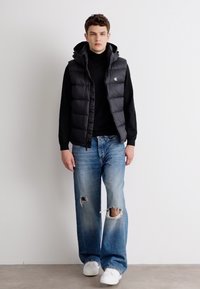 Zwarte puffer vest over een zwarte coltrui, gecombineerd met lichtblauwe distressed jeans en witte sneakers. Eenvoudig ontwerp en relaxte pasvorm.