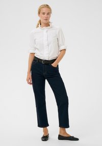 Weiße Bluse mit Rüschenturm, schwarzer Gürtel, dunkelblaue Jeans und schwarze Ballerinas. Die Jeans haben einen geraden Schnitt und dezente Nähte.