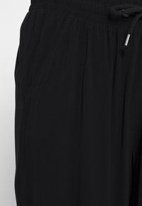 Pantalons noirs avec une taille élastique et un cordon de serrage, comprenant une poche latérale et un tissu léger et doux avec une texture plissée.