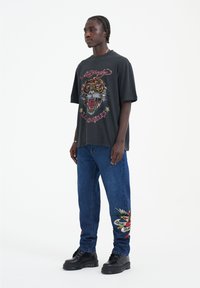 Camiseta negra de gran tamaño con un gráfico de tigre y texto. Pantalones vaqueros de denim azul con un diseño bordado en la pierna izquierda. Botas negras gruesas.