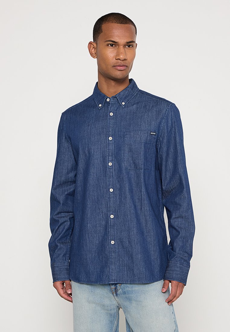 Homme portant une chemise en denim bleu foncé boutonnée avec une poche poitrine, associée à un jean bleu clair, se tenant devant un fond uni.