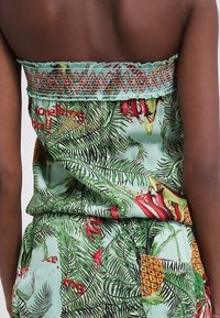 Femme portant une tenue bustier à imprimé tropical avec des feuilles vertes, des fleurs rouges et des motifs d'ananas, montrant son dos et ses bras nus.