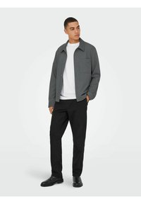 Graues Hemd mit Knopfleiste und einer Brusttasche, kombiniert mit schwarzen Hosen und schwarzen Stiefeln. Darunter wird ein schlichtes weißes T-Shirt getragen.