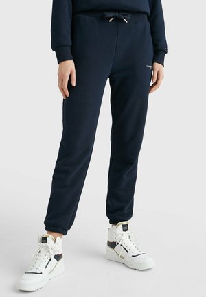 Persona que lleva pantalones joggers azul marino con puños elásticos y zapatillas blancas de caña alta con detalles en negro y dorado.