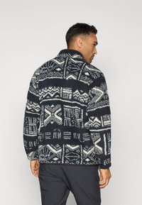 Fleece dzseki fekete és krém mintával, magas gallérral és laza fazonnal. Texturált anyag geometriai formákkal.