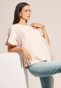 Helles pinkes Blusenshirt mit ausgestellten Ärmeln, glatte Textur. Kombiniert mit hellblauen Jeans, dazu eine gewebte weiße Tasche und Perlenaccessoires.