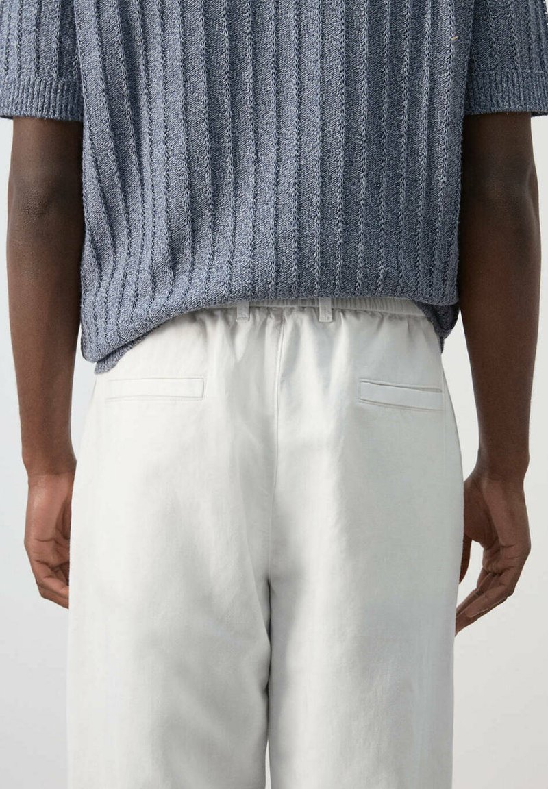 Personne portant un haut gris en maille texturée à manches courtes, rentré dans un pantalon blanc taille haute, vue de dos.