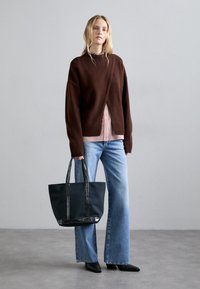 Un pull surdimensionné marron superposé sur une chemise rayée, associé à un jean large bleu clair. Sac fourre-tout noir avec des accents brillants.